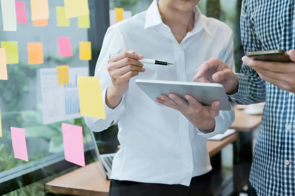 two men stand near the window, one is holding a tablet and a pen, other holds a phone