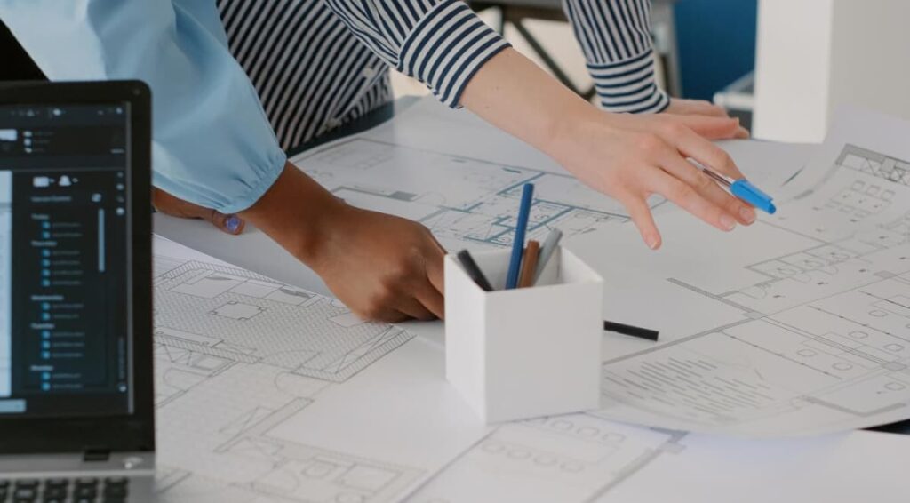 A person works on architectural plans with a computer nearby